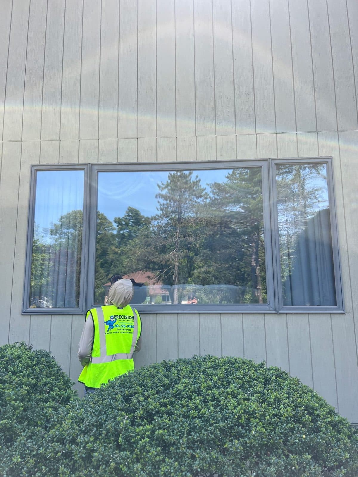 Precision Windows & Glass installer in a branded hi-vis vest finishing a large three-lite picture window with flanking units on a board-and-batten Passaic, NJ home